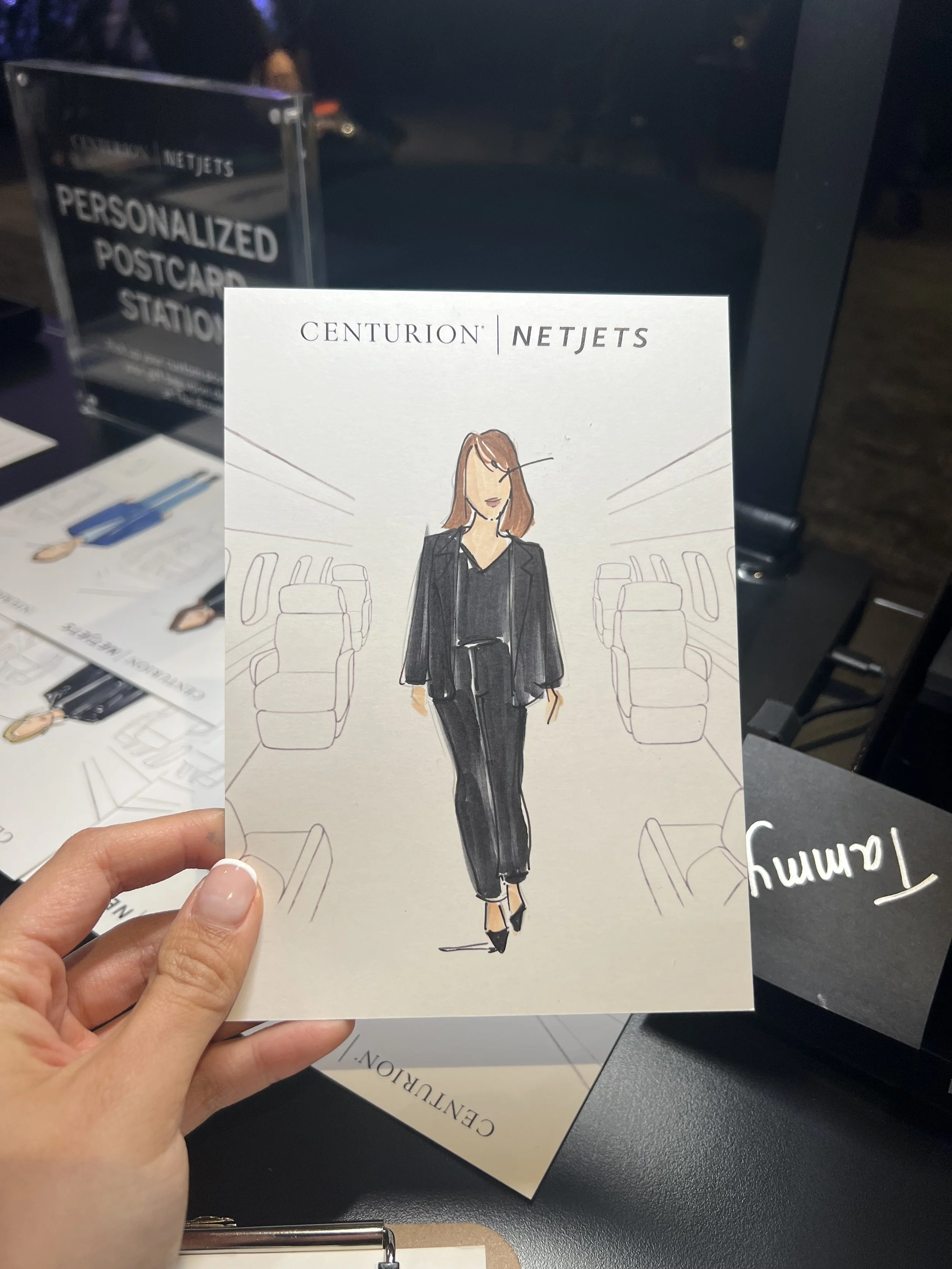 Hand holding a postcard with an illustration of a person in a black outfit inside an airplane cabin, featuring the text "Centurion NetJets". Background shows a personalized postcard station sign.
