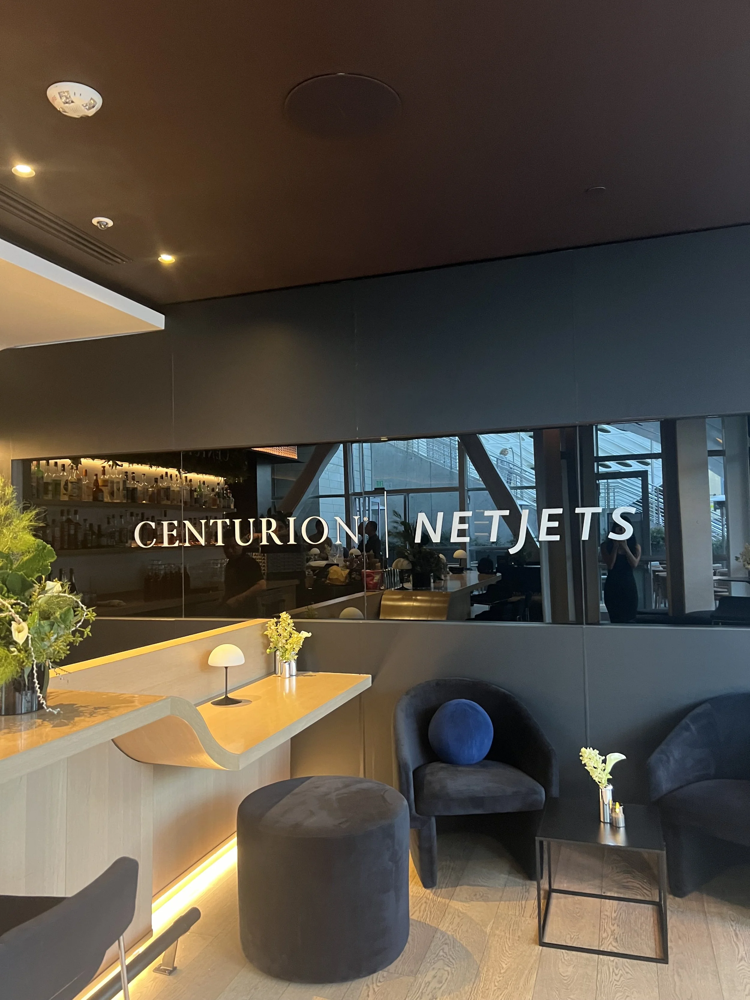 Lounge area with a sleek, modern design featuring black wall paneling with "Centurion NetJets" signage. The seating includes plush chairs and ottomans, with small tables decorated with flowers. A curved wooden counter is illuminated underneath, and a
