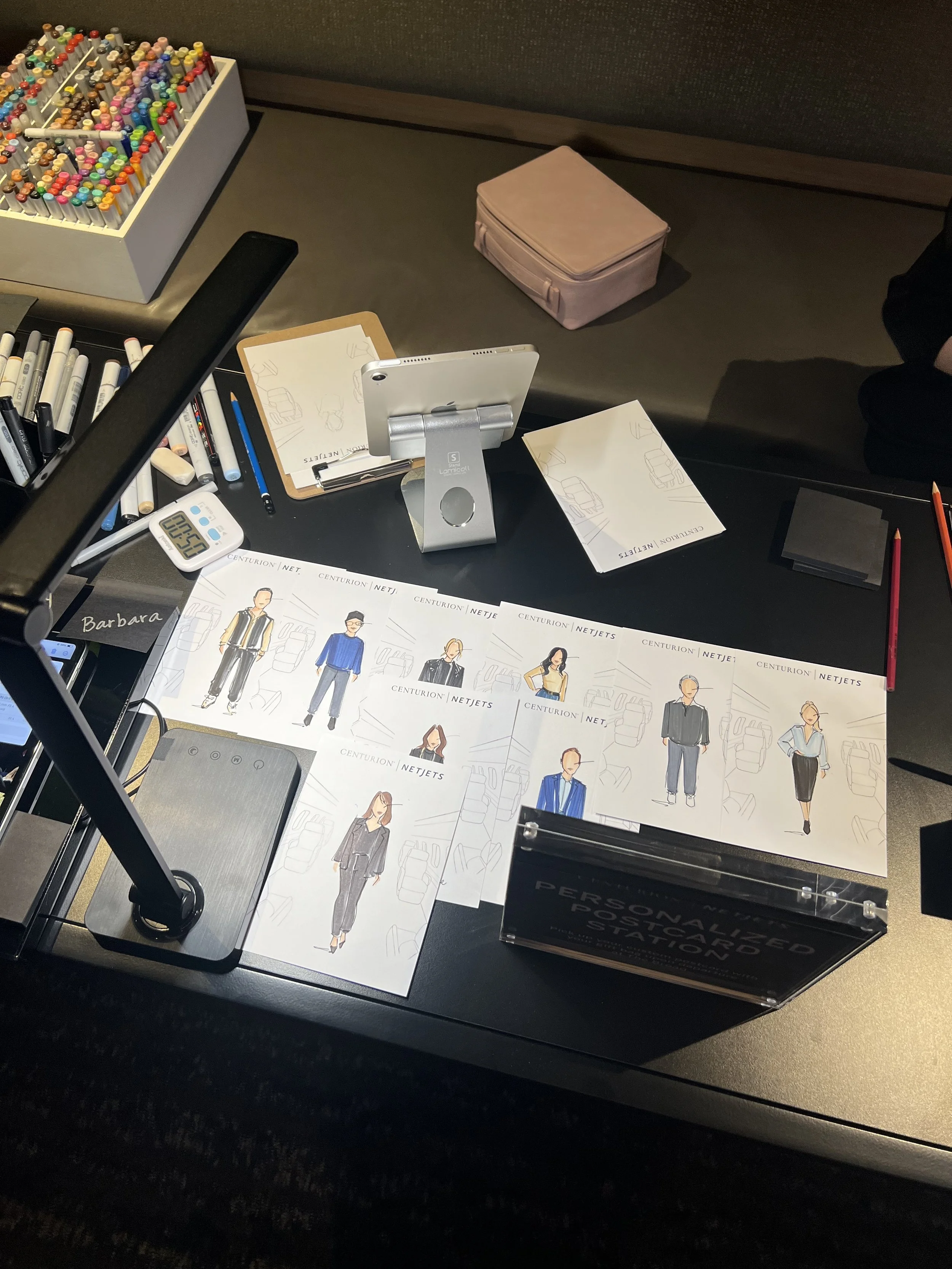 Table with personalized postcards, colorful pens, a tablet on a stand, and drawing supplies.