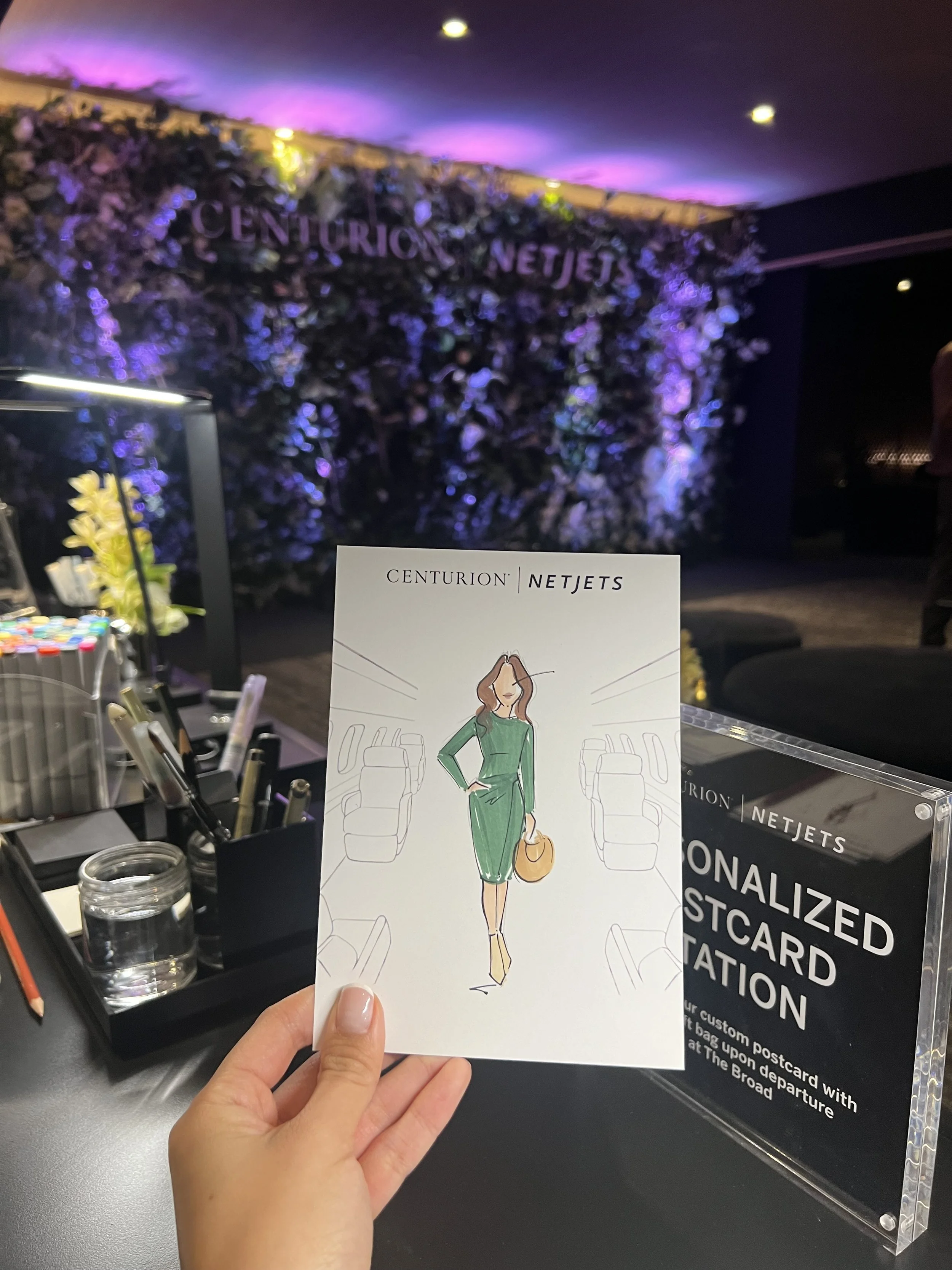 A hand holding a Centurion | NetJets postcard featuring an illustrated woman in a green dress. In the background, there's a desk with art supplies and personalized postcard station signage. The backdrop displays the Centurion NetJets logo with decora