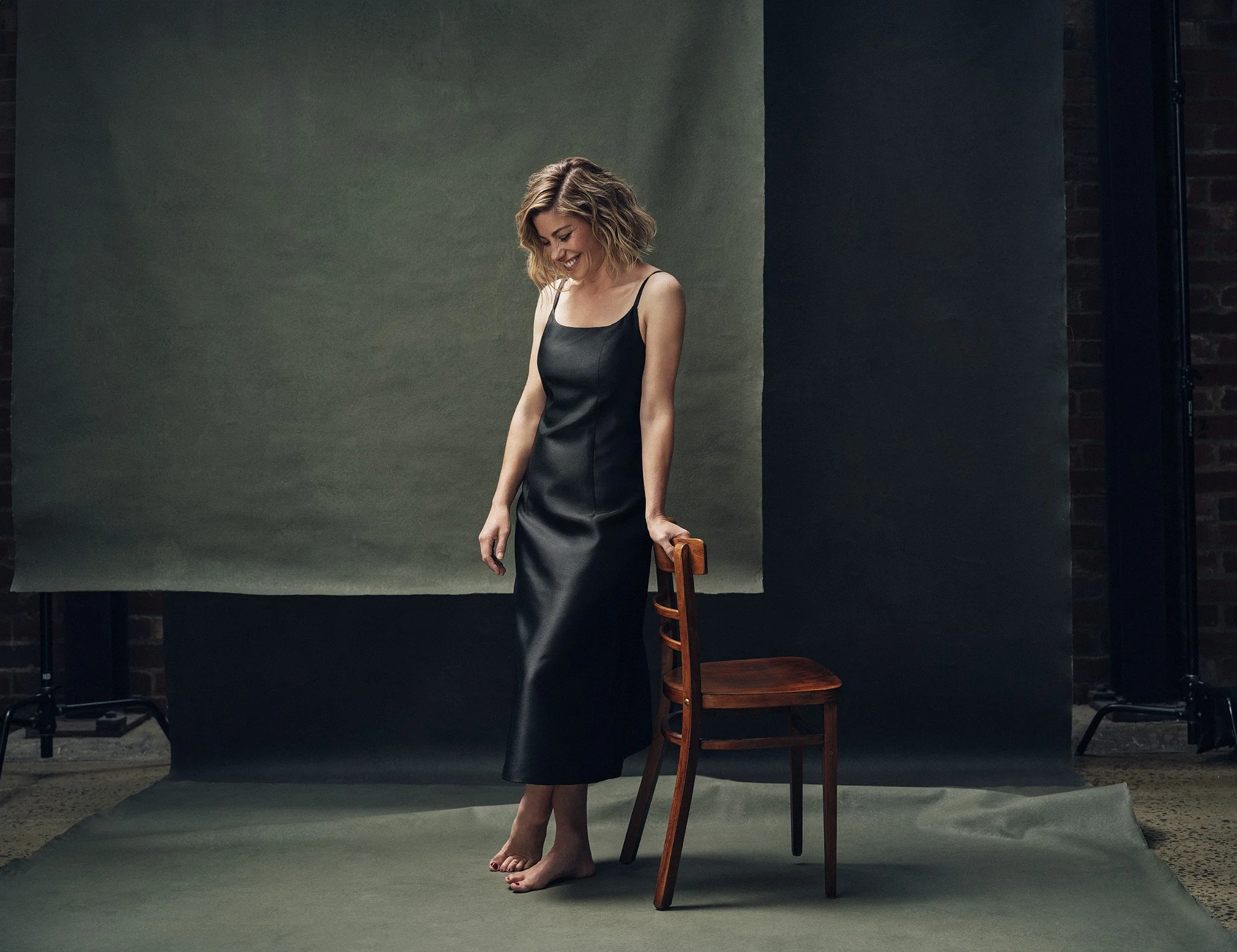 Brooke Satchwell posing in front of a canvas backdrop, smiling and leaning on chair with one hand