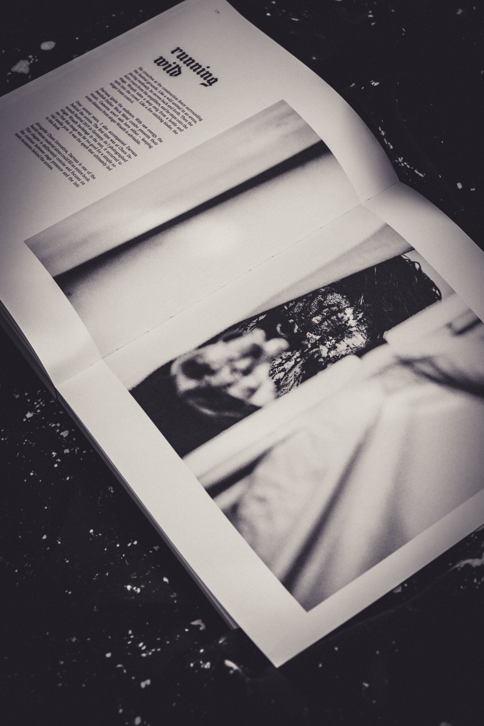 Schwarz-weiß Fotografie eines offenen Buches mit Text und Bildern, liegend auf einem dunklen, stark gesprenkelten Oberflächenhintergrund.