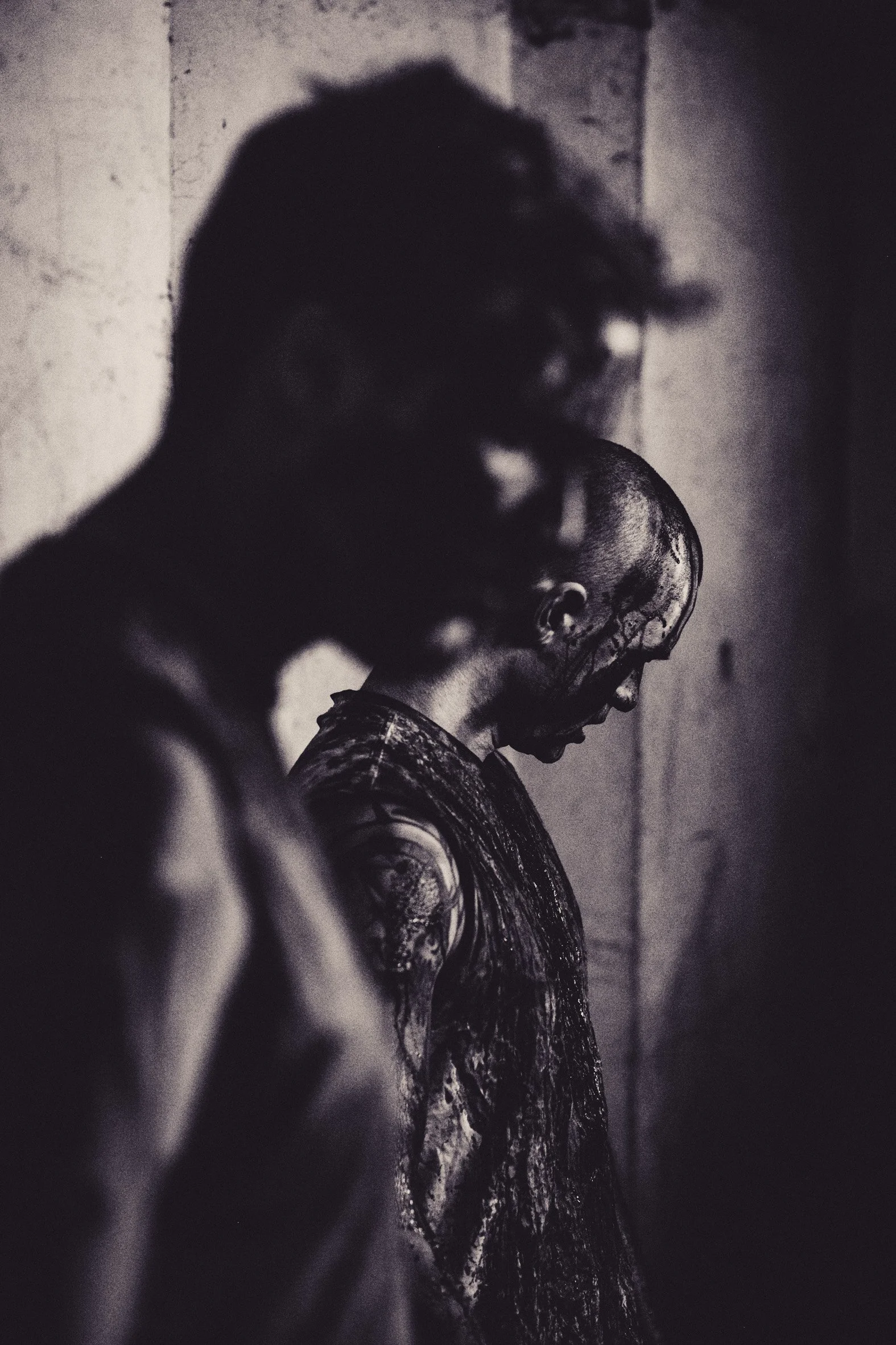 Schwarz-weiß-Foto von einer Person mit Tattoos, die den Kopf nach unten neigt, vor einer Steinmauer, mit betonter Schattierung und Stimmung.