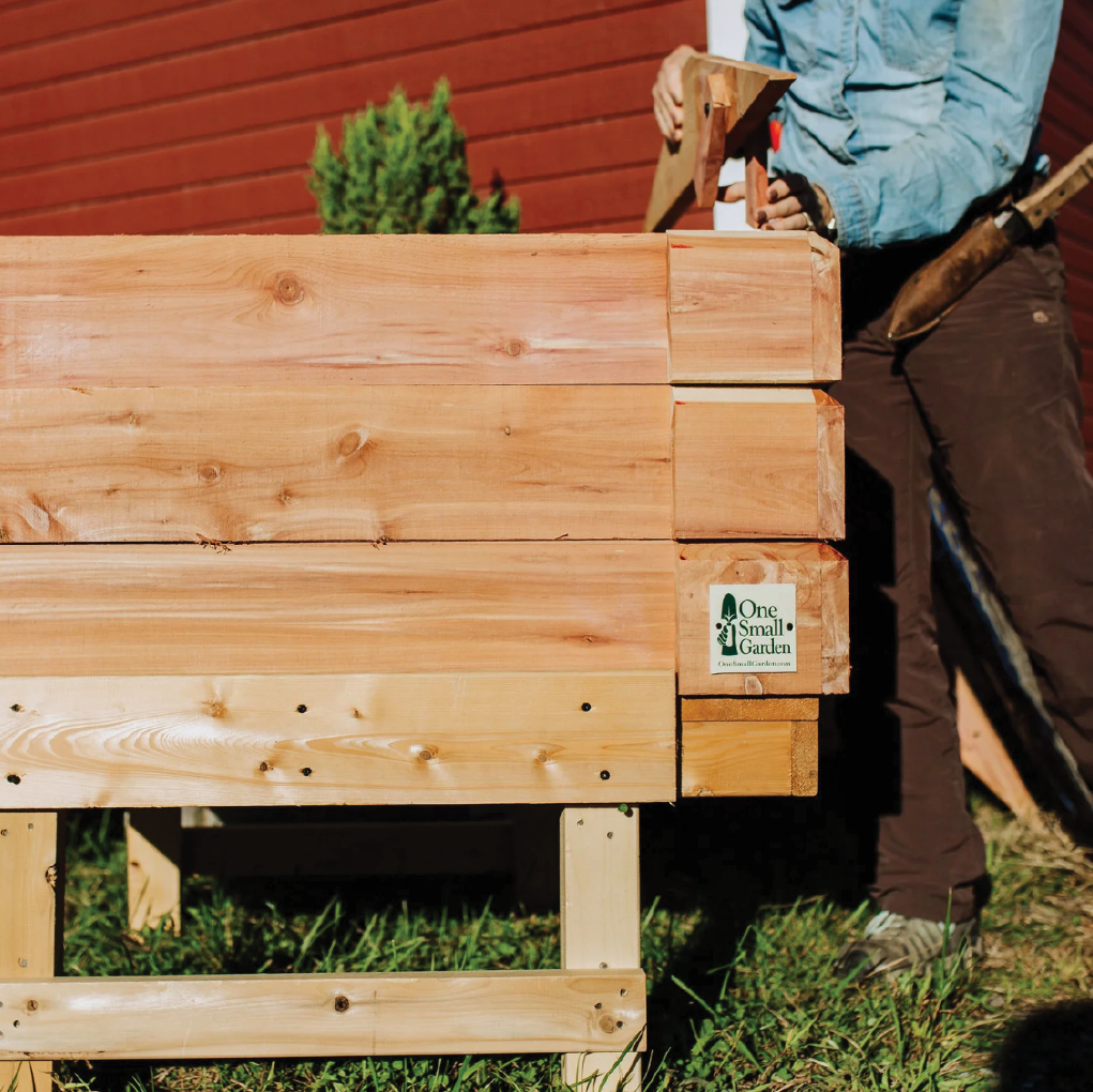 Standing garden bed made from cedar with One Small Garden logo on the front sits in the grass in front of a red building.  A gardener tends to the bed in the background.