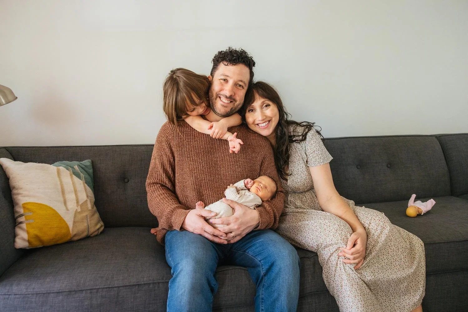 Family of four sitting together on a couch during an in-home newborn lifestyle session in Durham NC — parents, toddler girl, and sleeping newborn baby