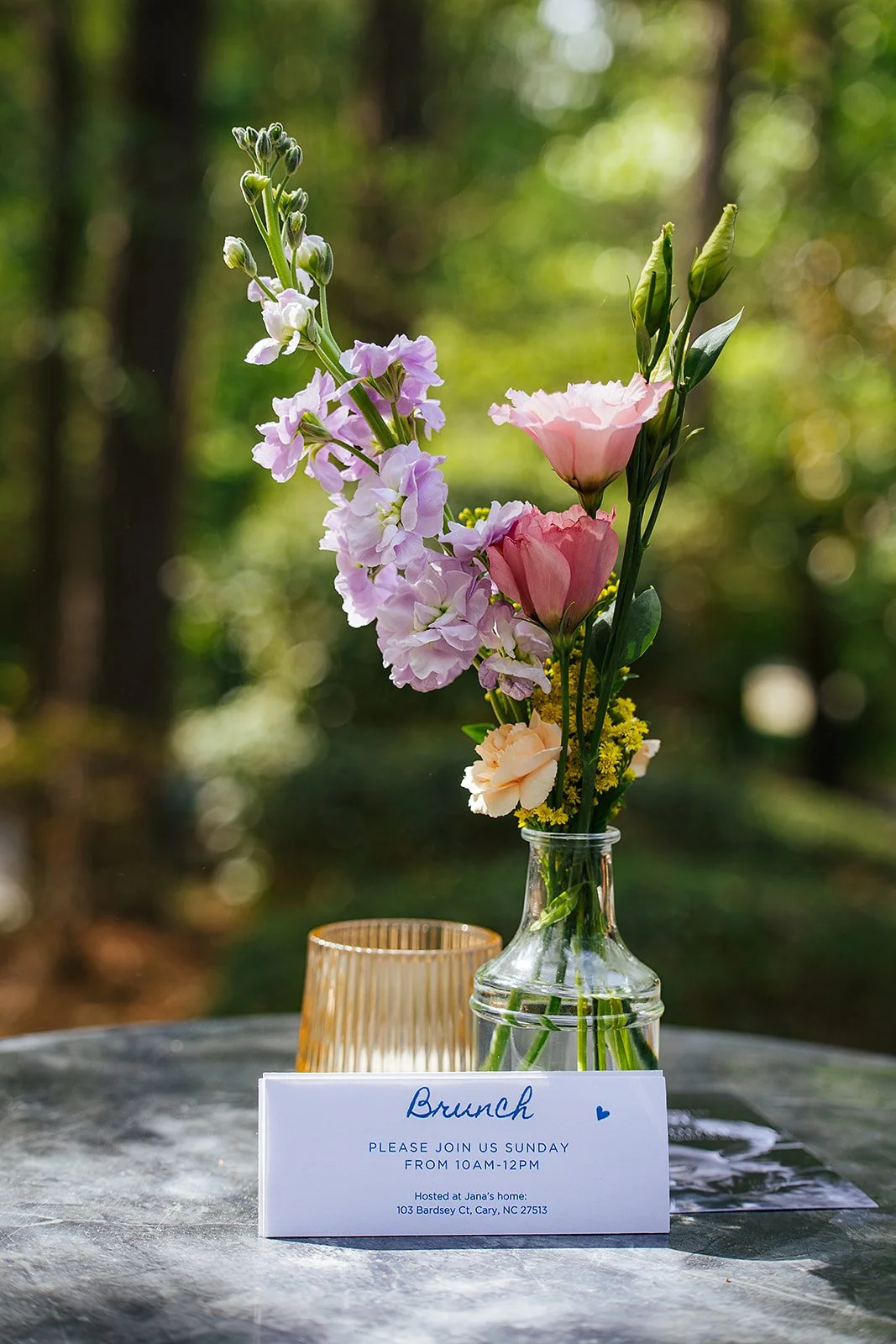 A glass bud vase with purple stock and pink lisianthus flowers sits beside a handwritten Brunch invitation card on a table, Durham NC wedding detail