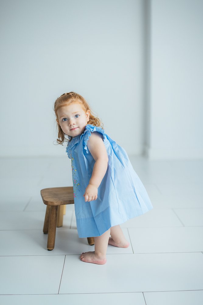 A young girl in a blue dress poses with a stool for a milestone portrait session in Durham, NC