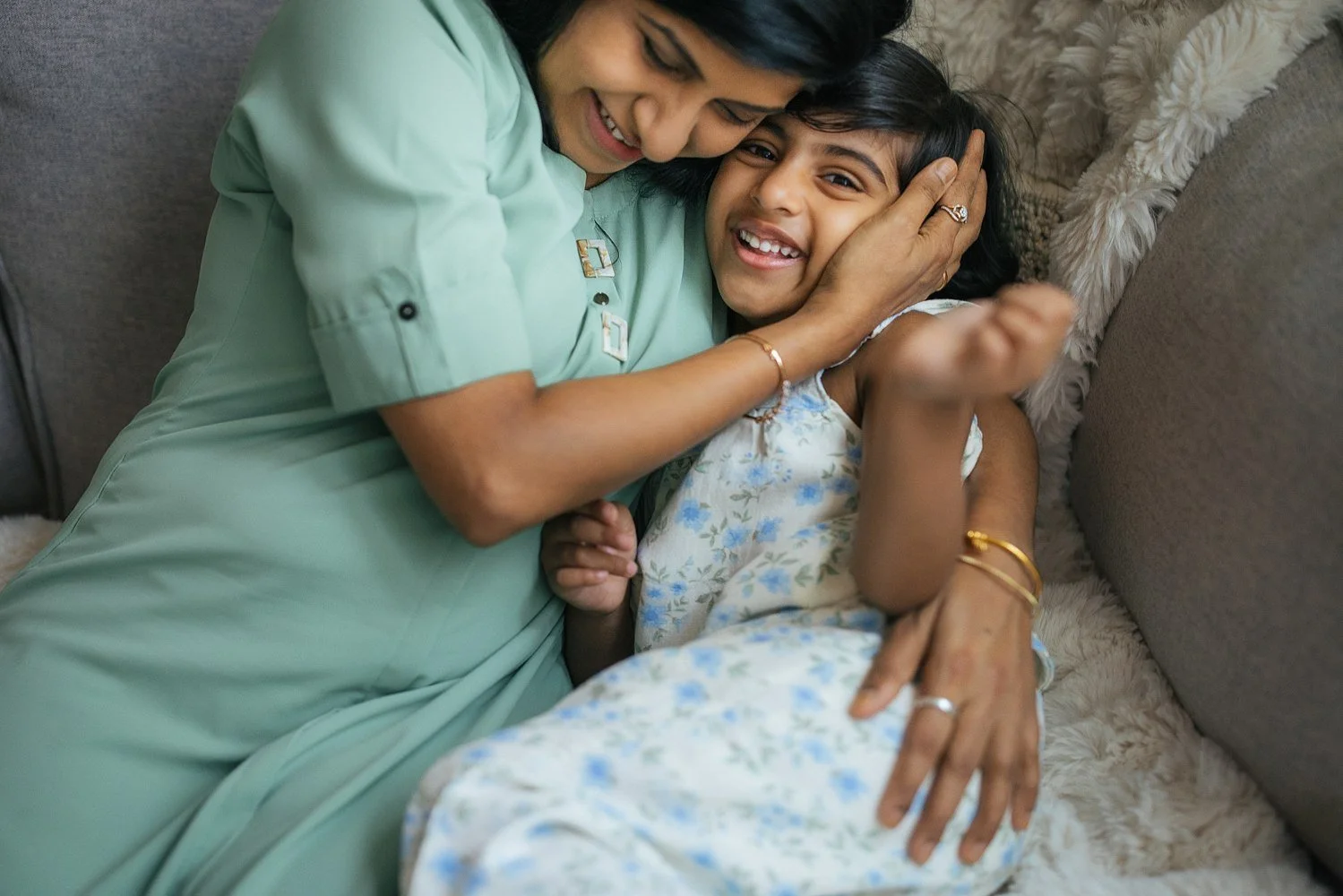 Mother in a sage green top holds her daughter's face gently in both hands as they laugh together on the sofa — candid in-home family session in Apex, NC.