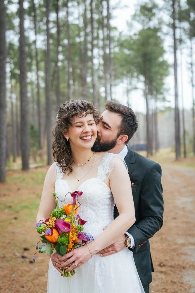 Bride Emily laughing with eyes closed while groom Connor kisses her cheek — colorful wildflower bouquet with orange calla lilies and purple blooms at Honeysuckle Tea House Chapel Hill NC