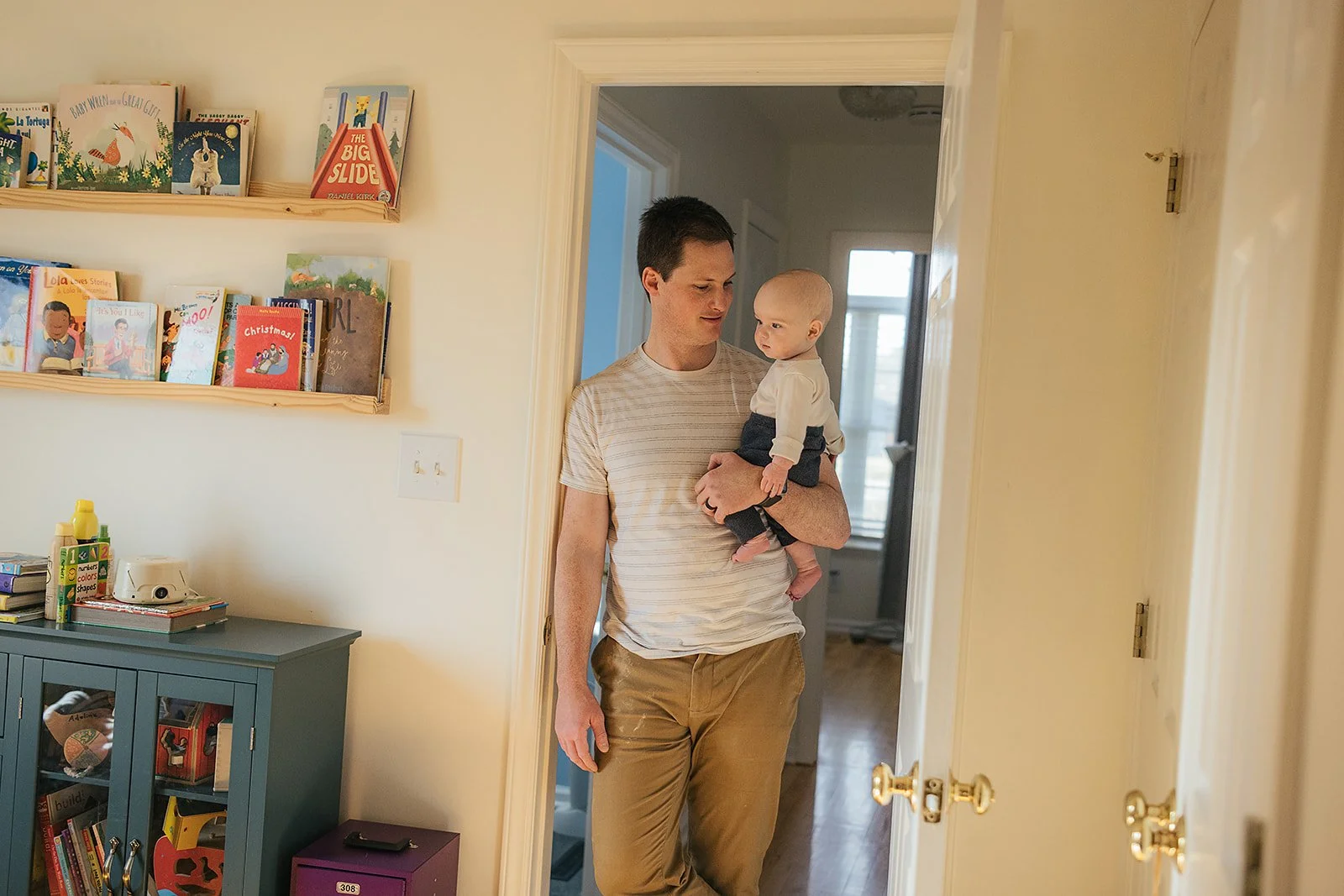 Dad carries baby into colorful children's room with bookshelves during Durham in-home family photography session