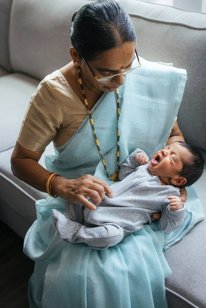 Grandmother in a pale blue saree sits on the sofa cradling her newborn grandson in both arms as the baby yawns widely with eyes closed — in-home newborn session in Apex, NC.