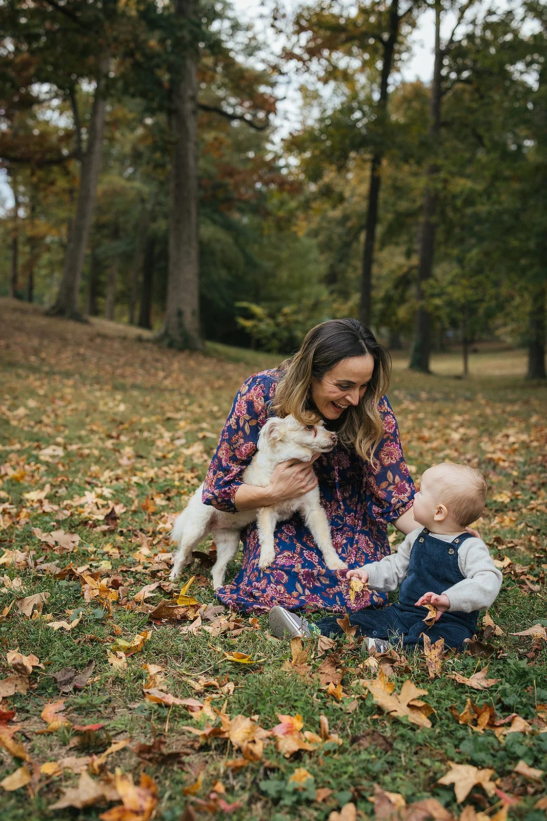 Baby boy reaches toward the family dog while mom kneels nearby in fall leaves at Duke Park during Durham NC motherhood photography session