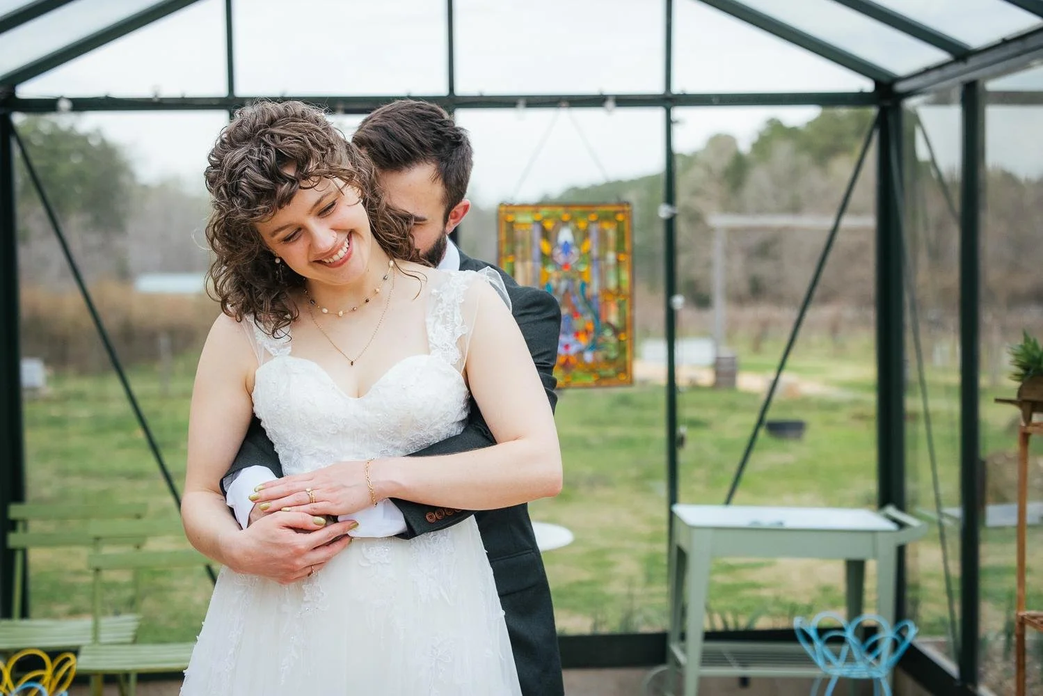 Groom holding bride from behind as she smiles and looks down — cozy intimate portrait inside the greenhouse at Honeysuckle Tea House Chapel Hill NC