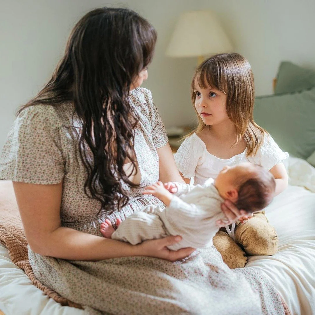 Mother holds a newborn while sitting on a bed with another child during an in home photo session in Durham, NC