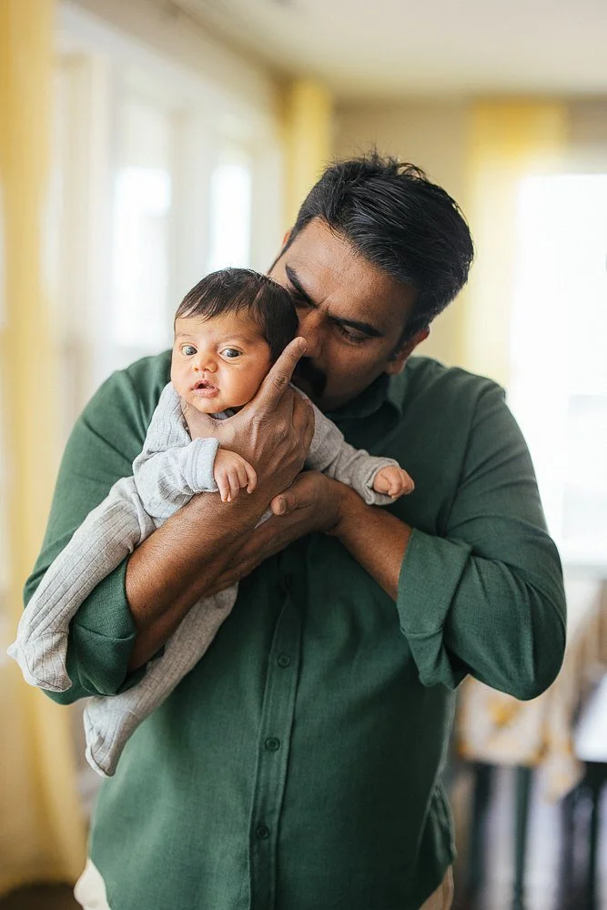 Father in a dark green linen shirt holds his newborn son up and leans in to kiss him tenderly near a bright window with yellow curtains — in-home newborn family session in Apex, NC.