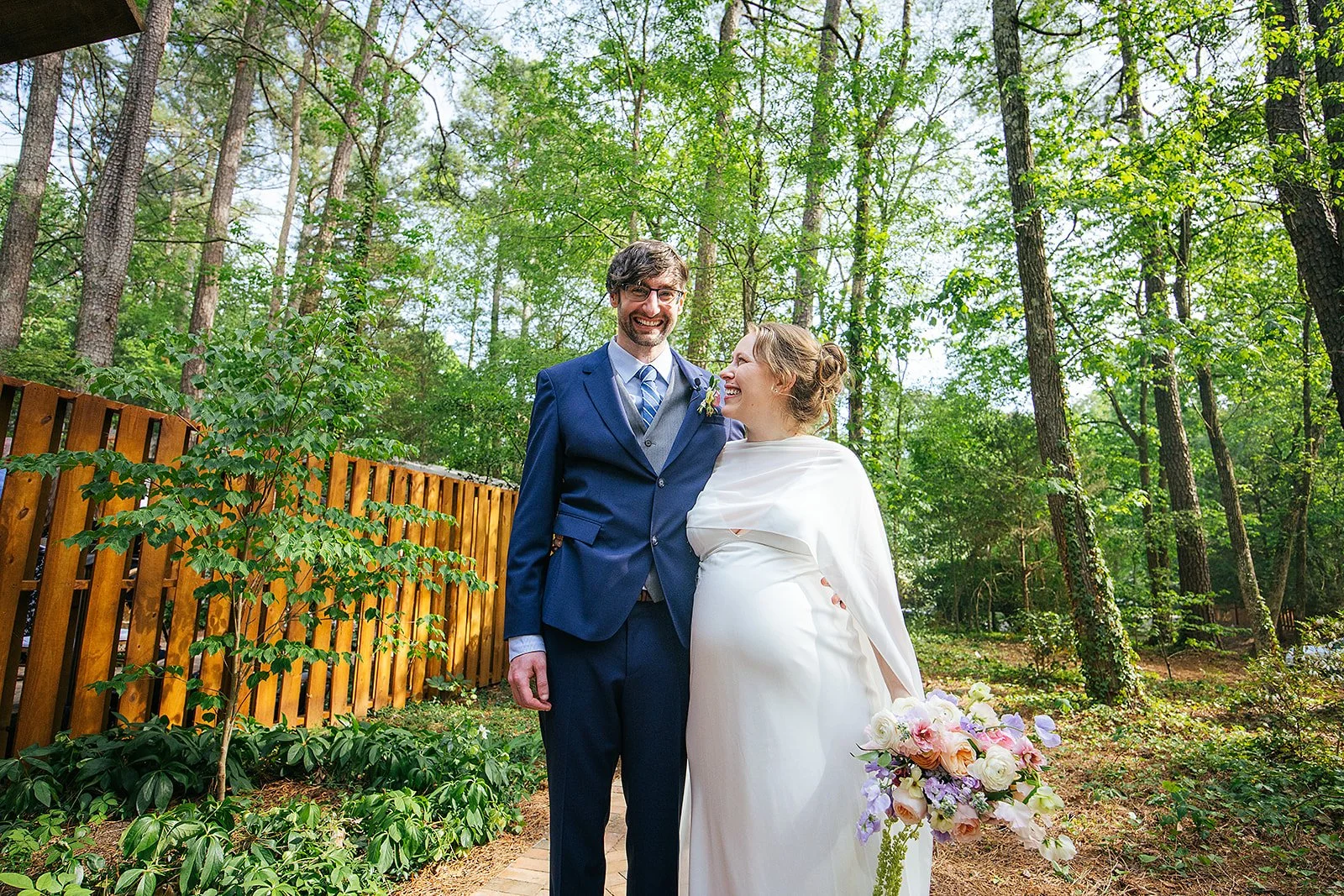 Newlywed couple laughs together on a wooded path with sunlight filtering through the trees, bride holding her spring bouquet, Durham NC wedding portraits