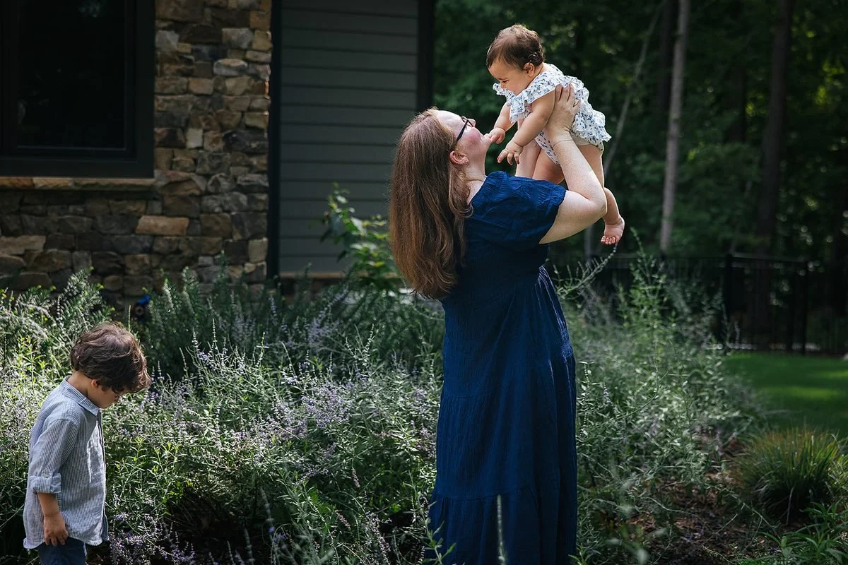 Mom holding baby in back yard while toddler son walks around