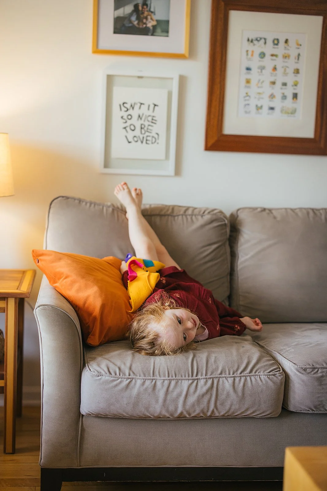 Toddler girl relaxes on couch in family living room with art print on wall during Durham lifestyle family photography session