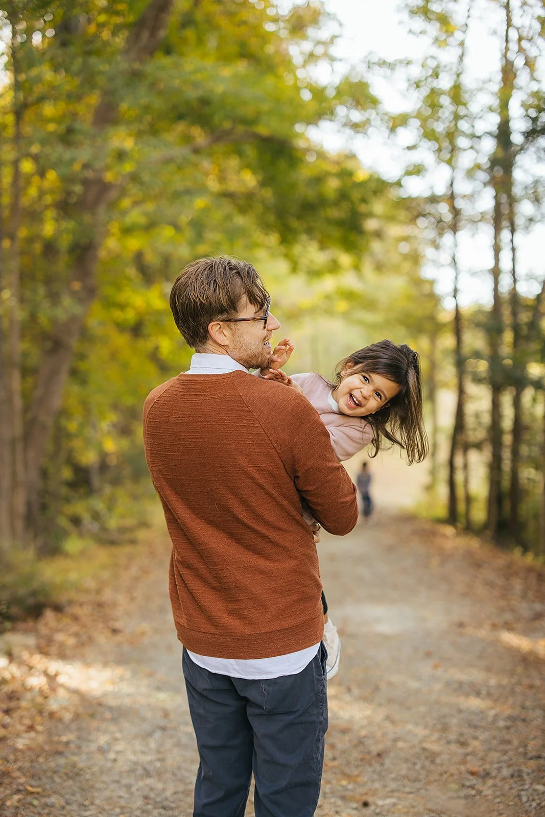 Dad lifting and spinning his laughing little girl on a tree-lined path at Blackwood Farm Park, surrounded by vibrant fall foliage during a Chapel Hill family session
