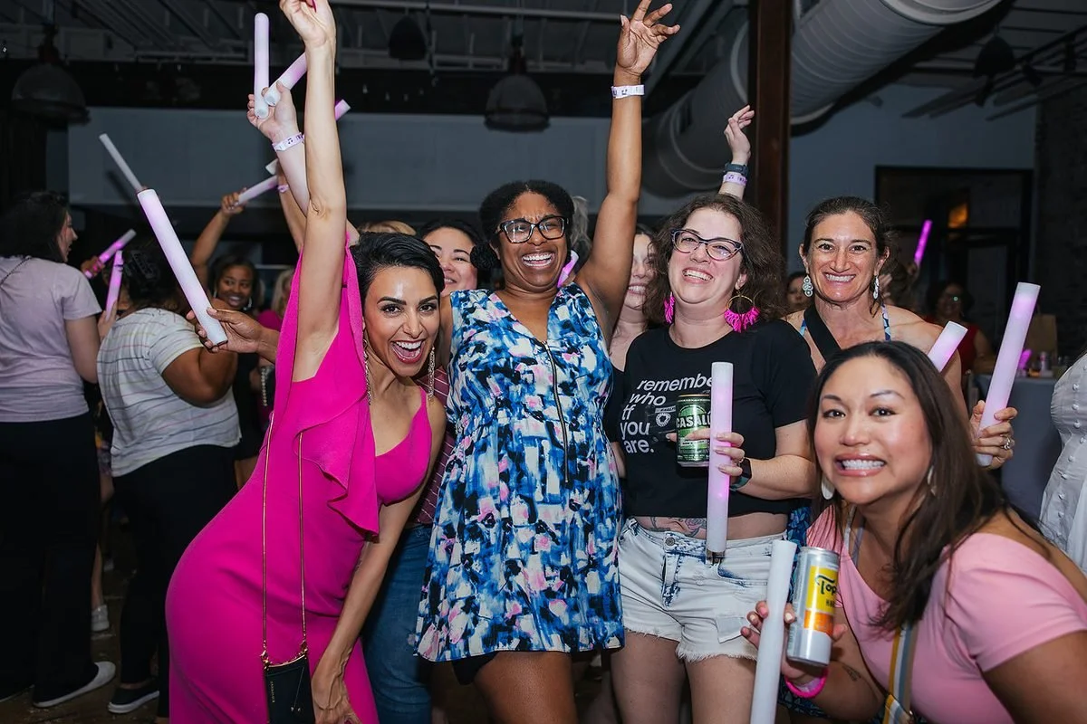 Group of five diverse women cheering together with glow sticks at the Dance Her popup in Raleigh — arms up, big smiles, pure joy