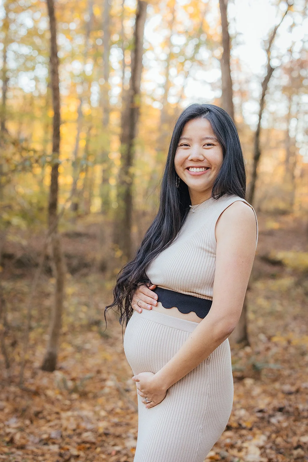A pregnant woman smiles in a wooded area for Fall family photos in Durham, NC