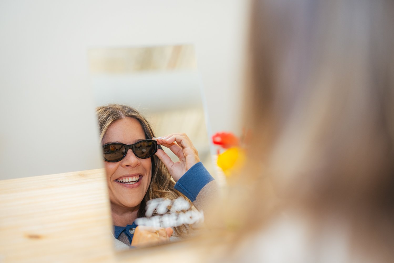 Mirror reflection of a smiling woman trying on sunglasses at a fun activity station at Longfellow Triangle LIFE corporate event in Raleigh NC