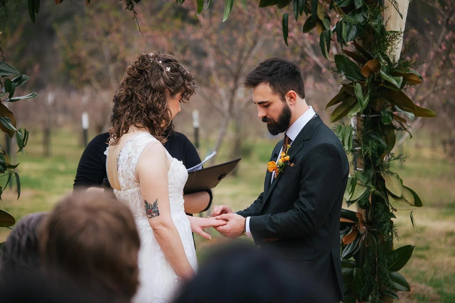 Bride placing a ring on groom's finger during the ring exchange ceremony under the magnolia arch at Honeysuckle Tea House Chapel Hill NC — officiant beside them