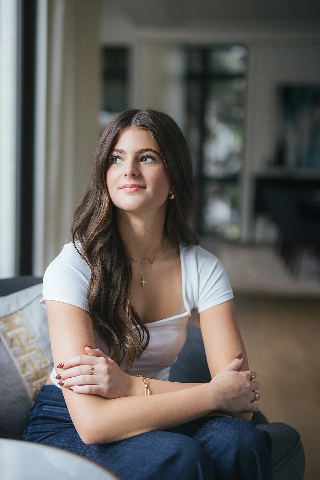 A teenage girl in a white top gazes softly upward while seated near a window, her expression quiet and reflective, in a portrait from her family's in-home session in downtown Raleigh, NC.