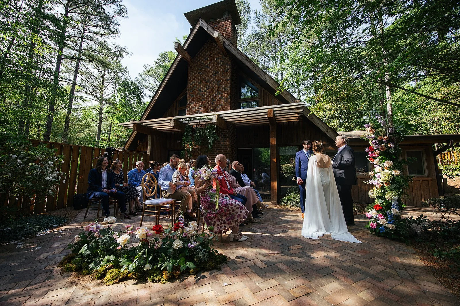 Wide view of an intimate backyard wedding ceremony with a small group of guests seated on gold chairs as the couple exchanges vows before a rustic wooden cabin, Durham NC