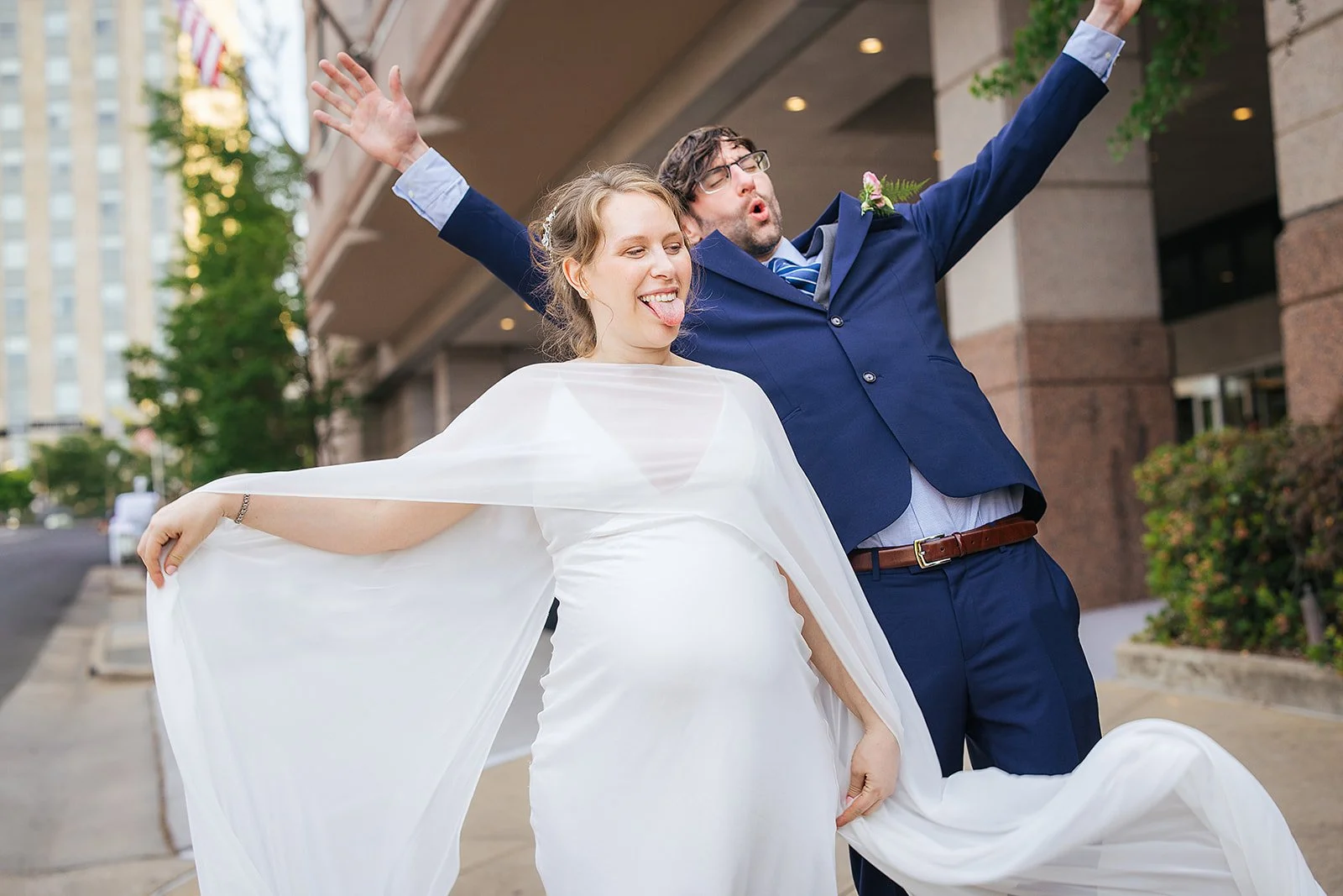 Pregnant bride spreads her white cape wide on a downtown Durham NC sidewalk with her tongue out playfully while groom throws his arms up in celebration behind her