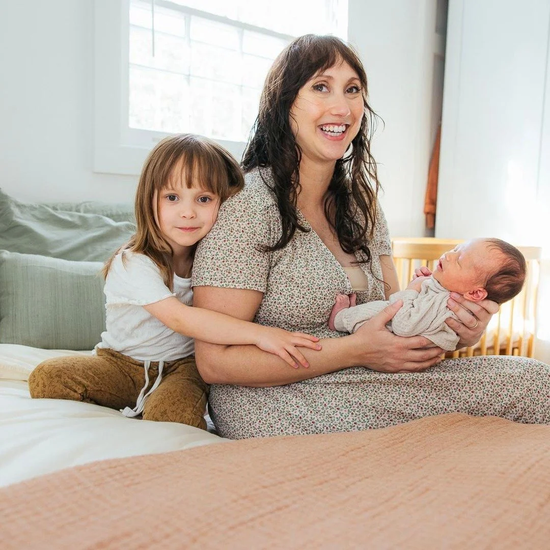 Mother smiles with a newborn and an older sibling during an at home photoshoot in Durham NC