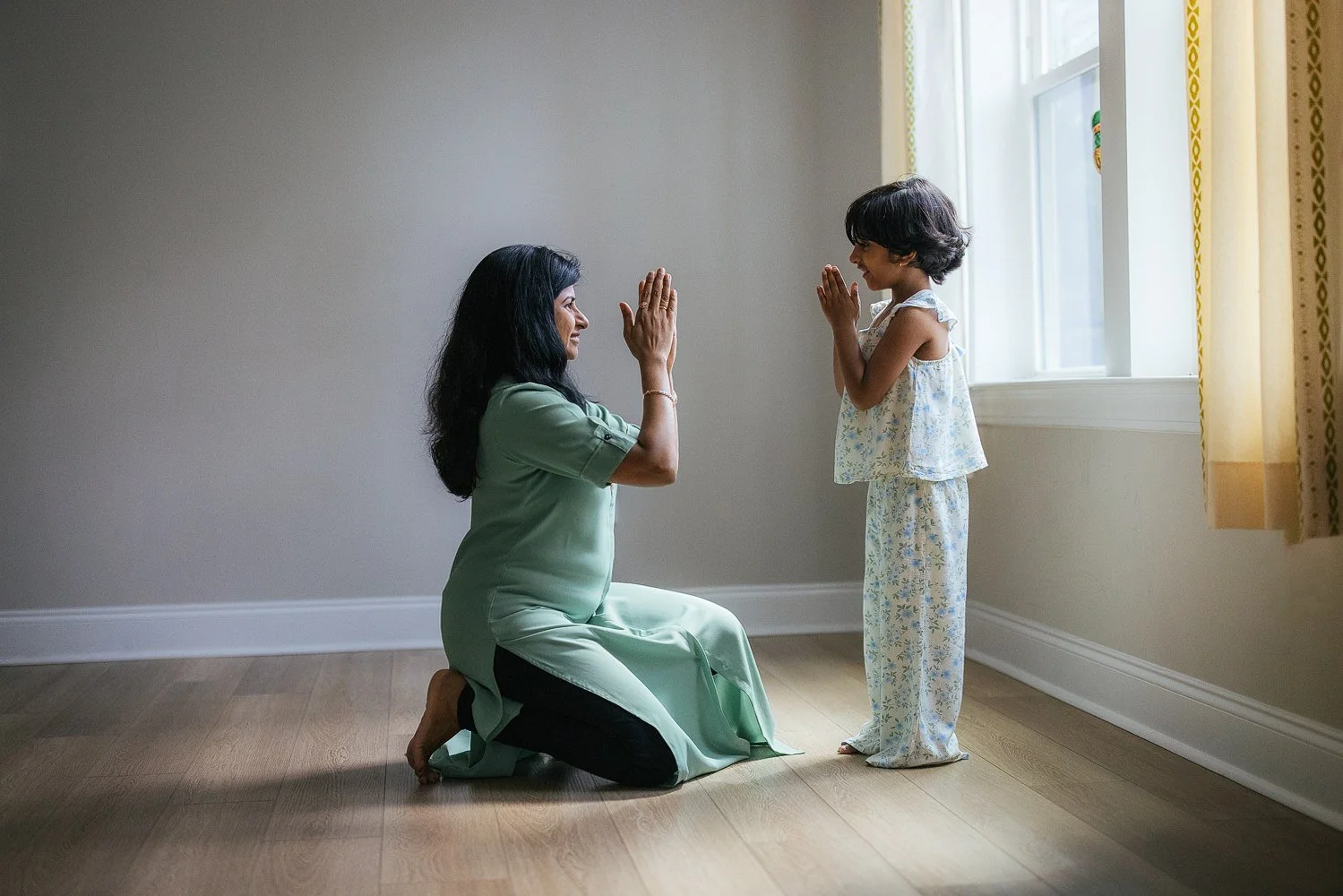 Mother and daughter face each other with palms pressed together in a classical dance gesture by a bright window — warm natural light fills the room — in-home family session in Apex, NC.