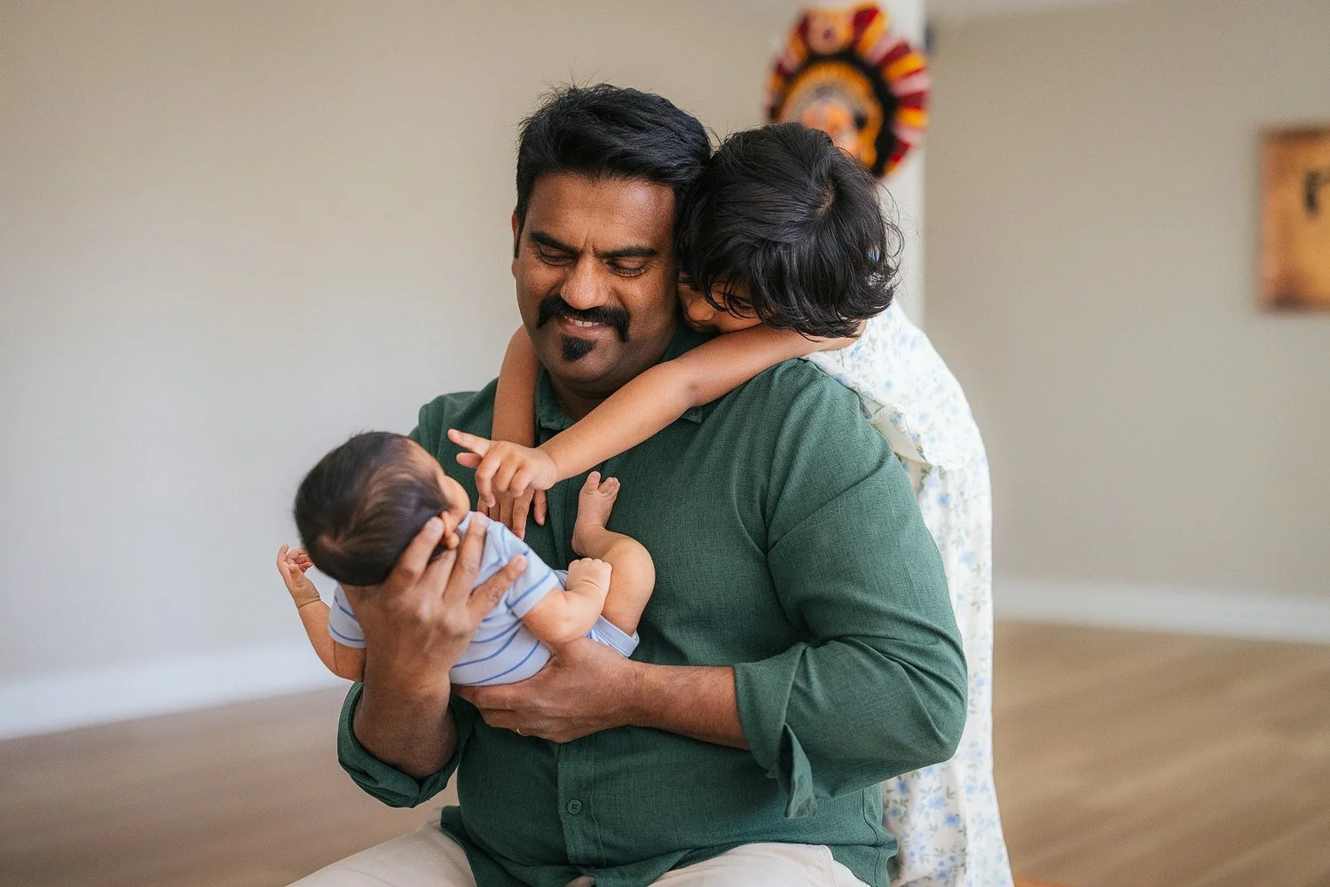 Father holds his newborn in both arms as his older daughter hugs him from behind with a wide grin — Kathakali mask visible on the wall — candid in-home session in Apex, NC.