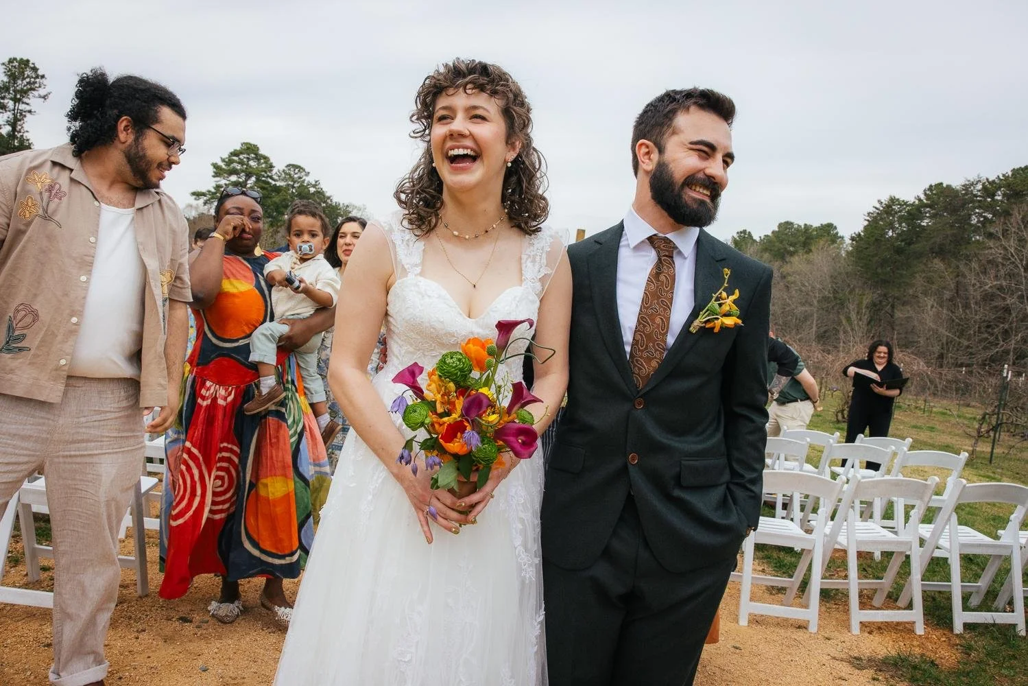 Bride and groom walking their recessional with joyful guests cheering in colorful outfits at the outdoor ceremony at Honeysuckle Tea House Chapel Hill NC