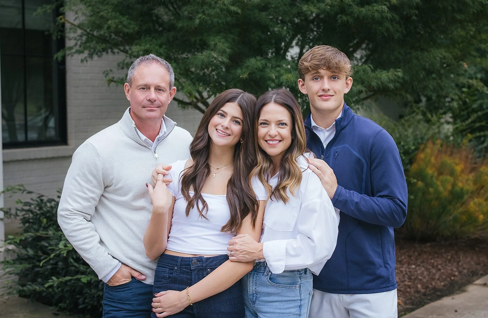 A family of four poses together outdoors among lush greenery near their home in downtown Raleigh, NC, smiling easily in a warm, relaxed group portrait.