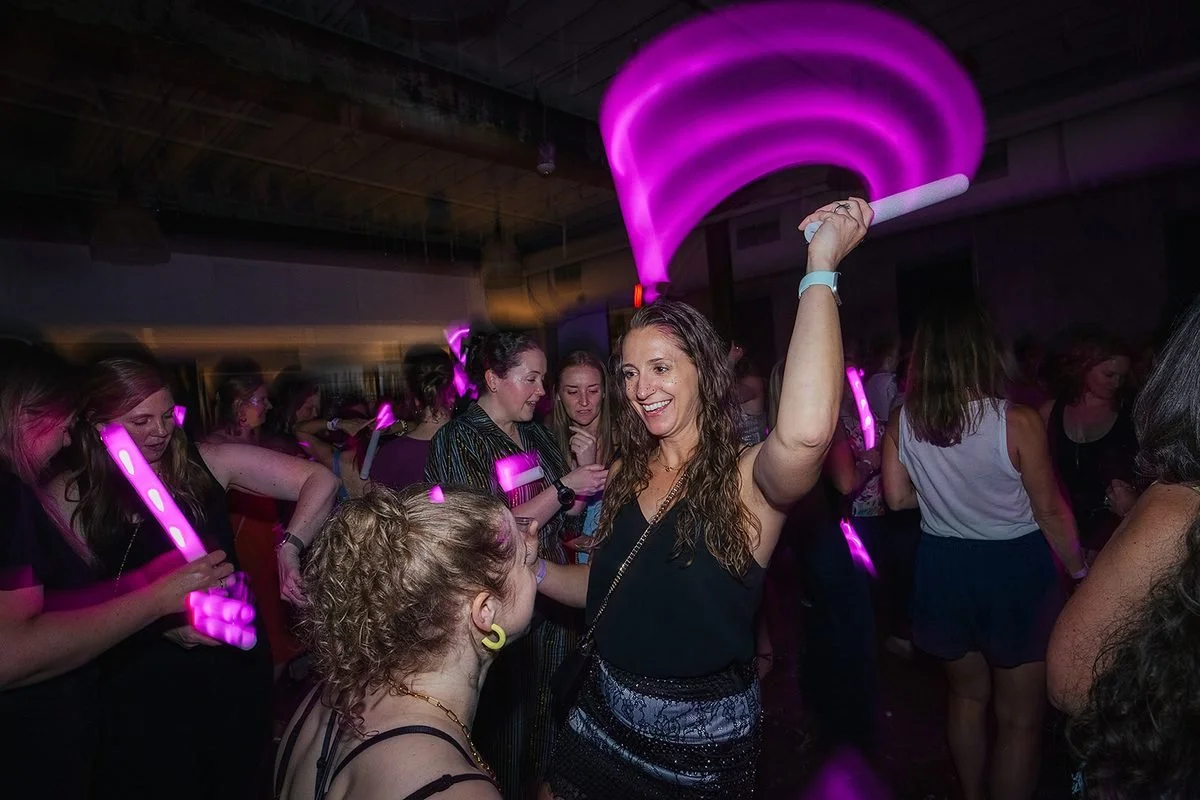 Woman waving a glowing pink foam stick above her head with a huge smile, surrounded by other women at the Dance Her popup in Raleigh