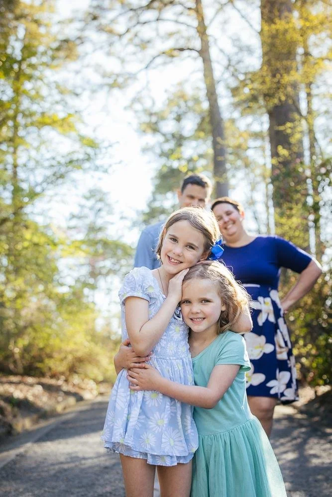 Two sisters hugging on a sunlit wooded path at Duke Gardens with parents softly blurred in background — Durham NC family photographer