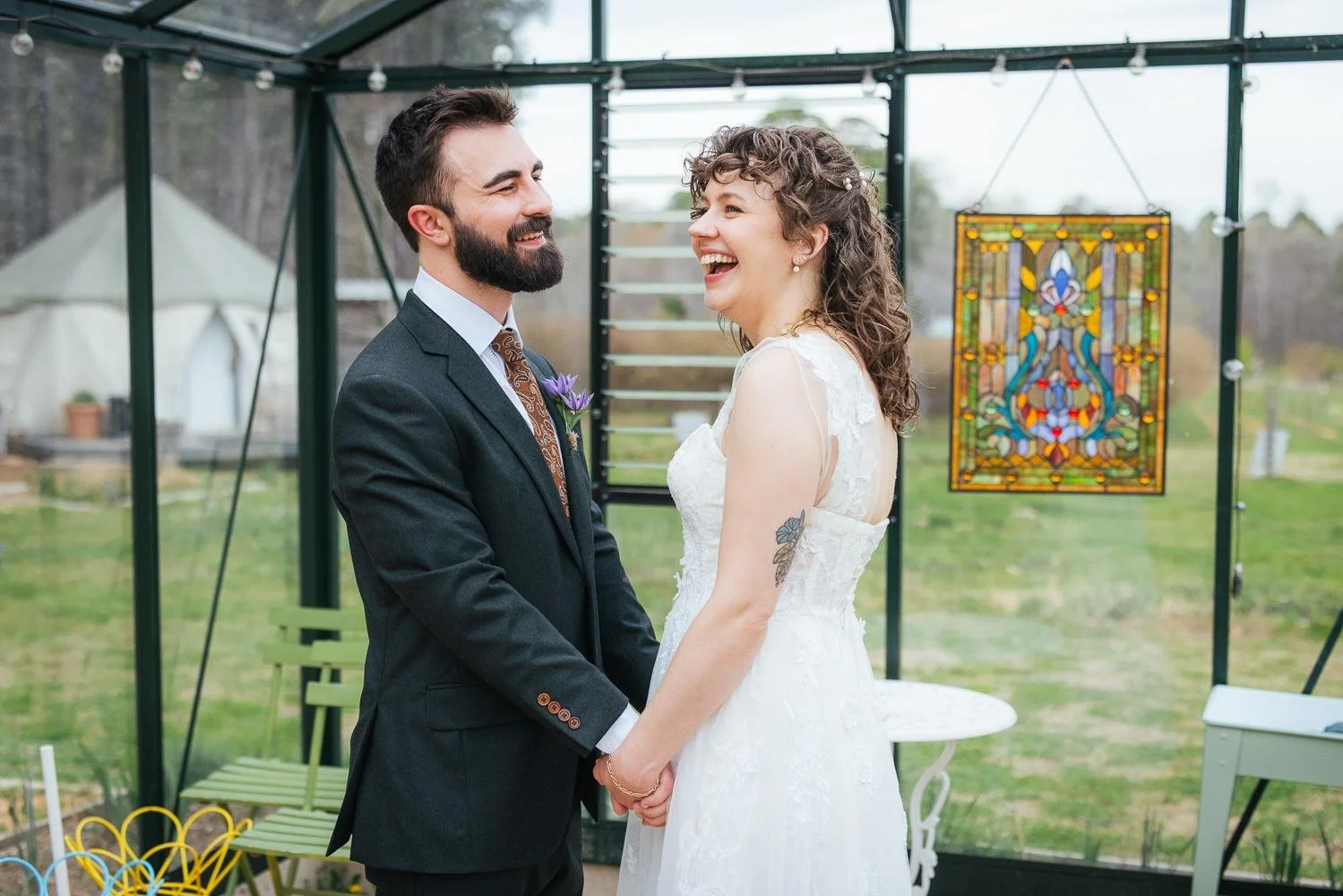Bride and groom laughing together inside the greenhouse at Honeysuckle Tea House — colorful stained glass window visible behind them — Chapel Hill NC wedding photographer