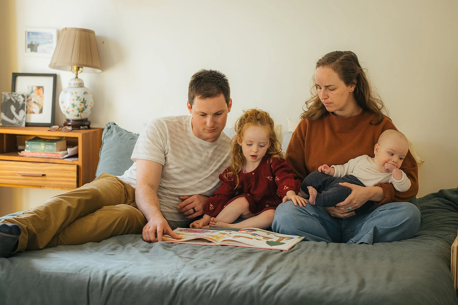 Mom, dad, toddler, and baby share storytime together on bed during Durham in-home family photography session