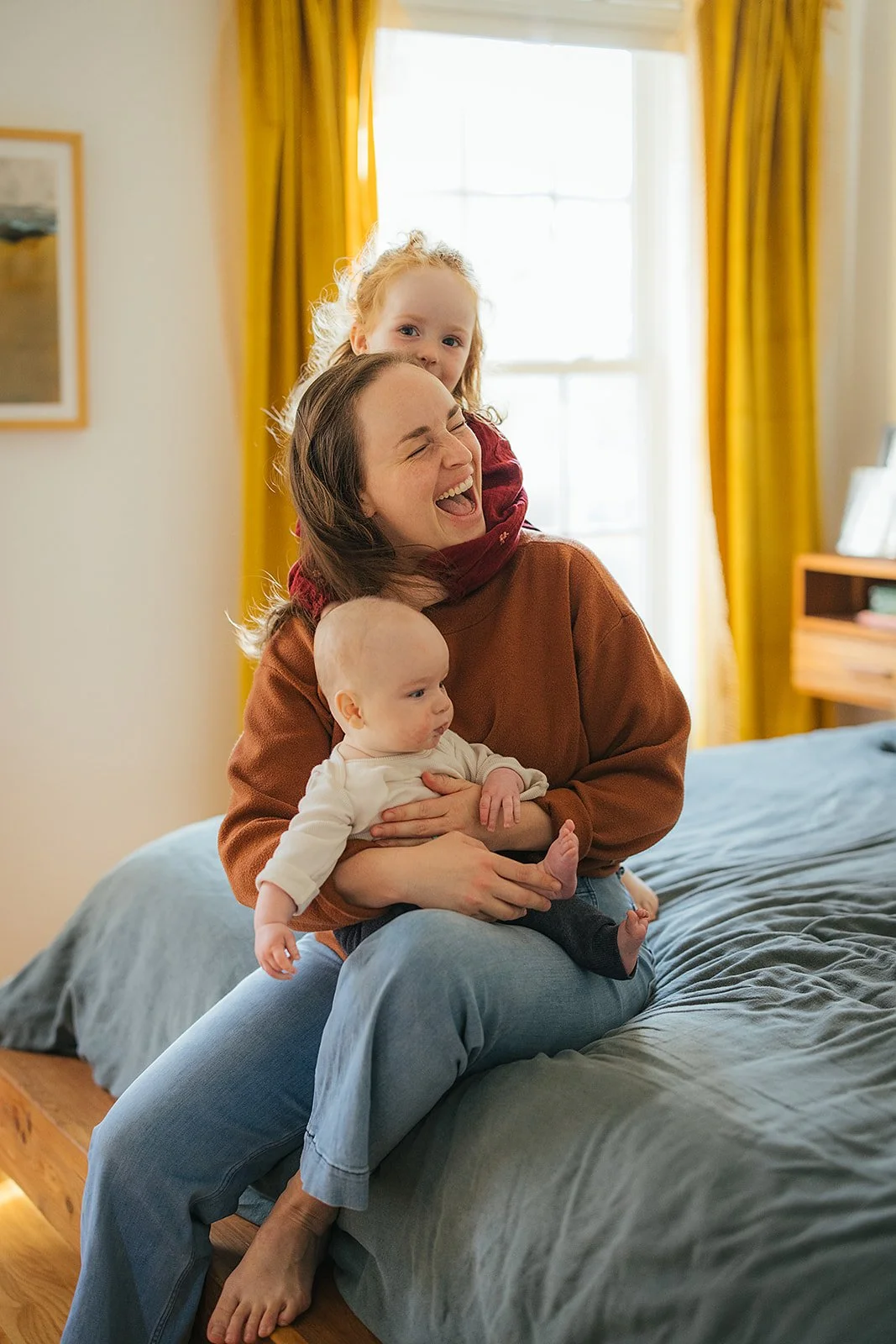 Mom laughs as toddler hugs her from behind while she holds baby during Durham in-home family photography session