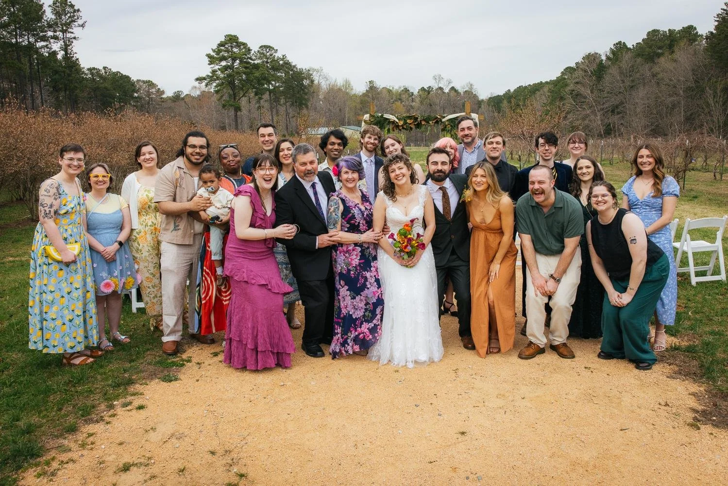 Full wedding group photo on the lawn at Honeysuckle Tea House in Chapel Hill NC — colorful guests surrounding the bride and groom on the sandy path