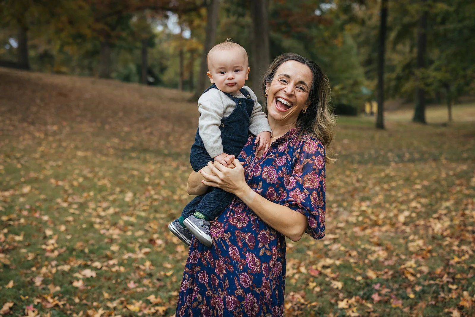 Mom laughs while holding her one - year - old baby boy in an open field at Duke Park during Durham NC motherhood photography session