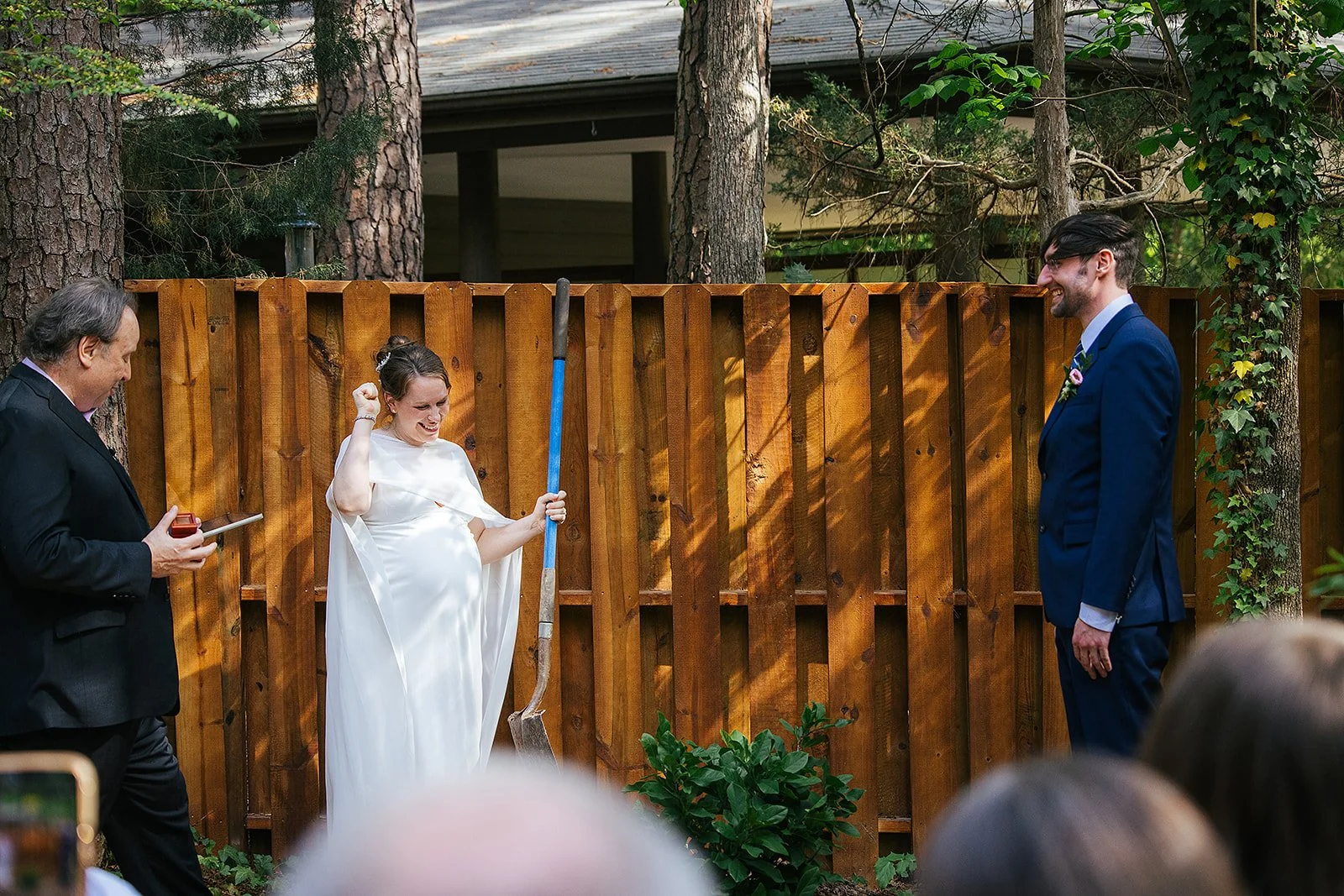Pregnant bride triumphantly pumps her fist mid-jump over a broom during a broom jumping ceremony tradition while her groom watches and laughs, Durham NC wedding