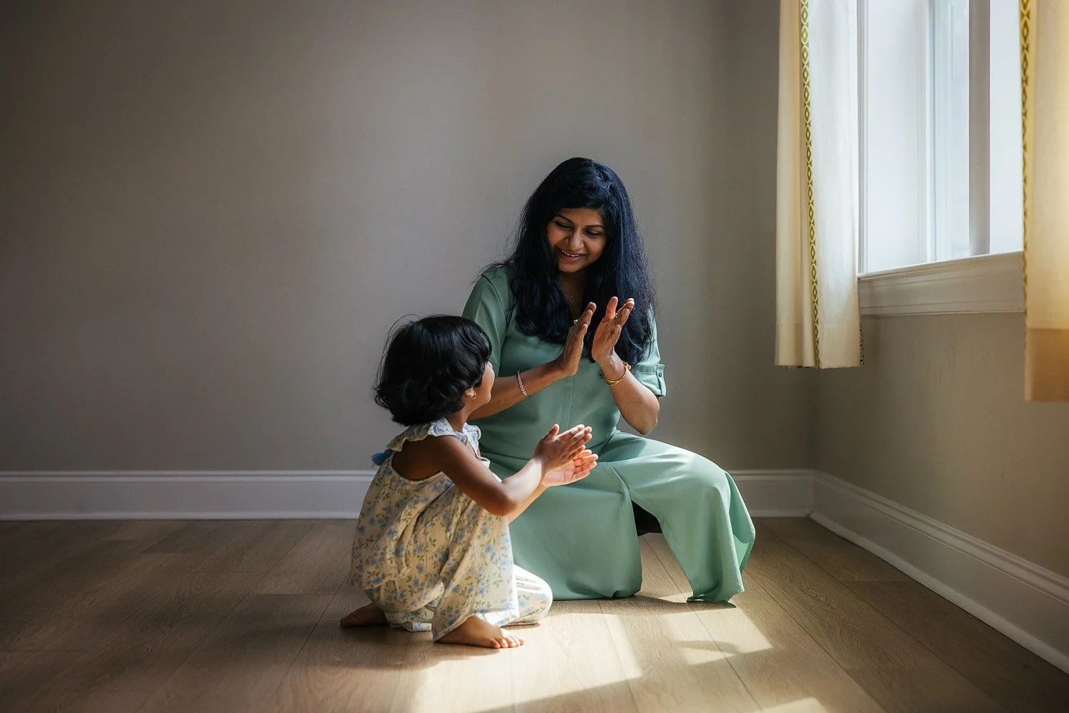 Mother and toddler daughter sit facing each other on hardwood floors playing a clapping game — soft natural window light fills the room — candid in-home family session in Apex, NC.