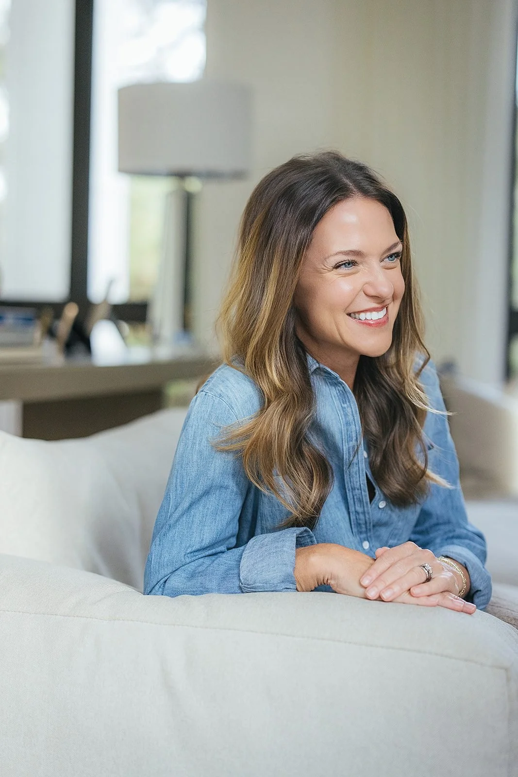 A mother in a soft denim shirt leans over the back of a cream sofa and smiles warmly, photographed during her family's in-home session in downtown Raleigh, NC.