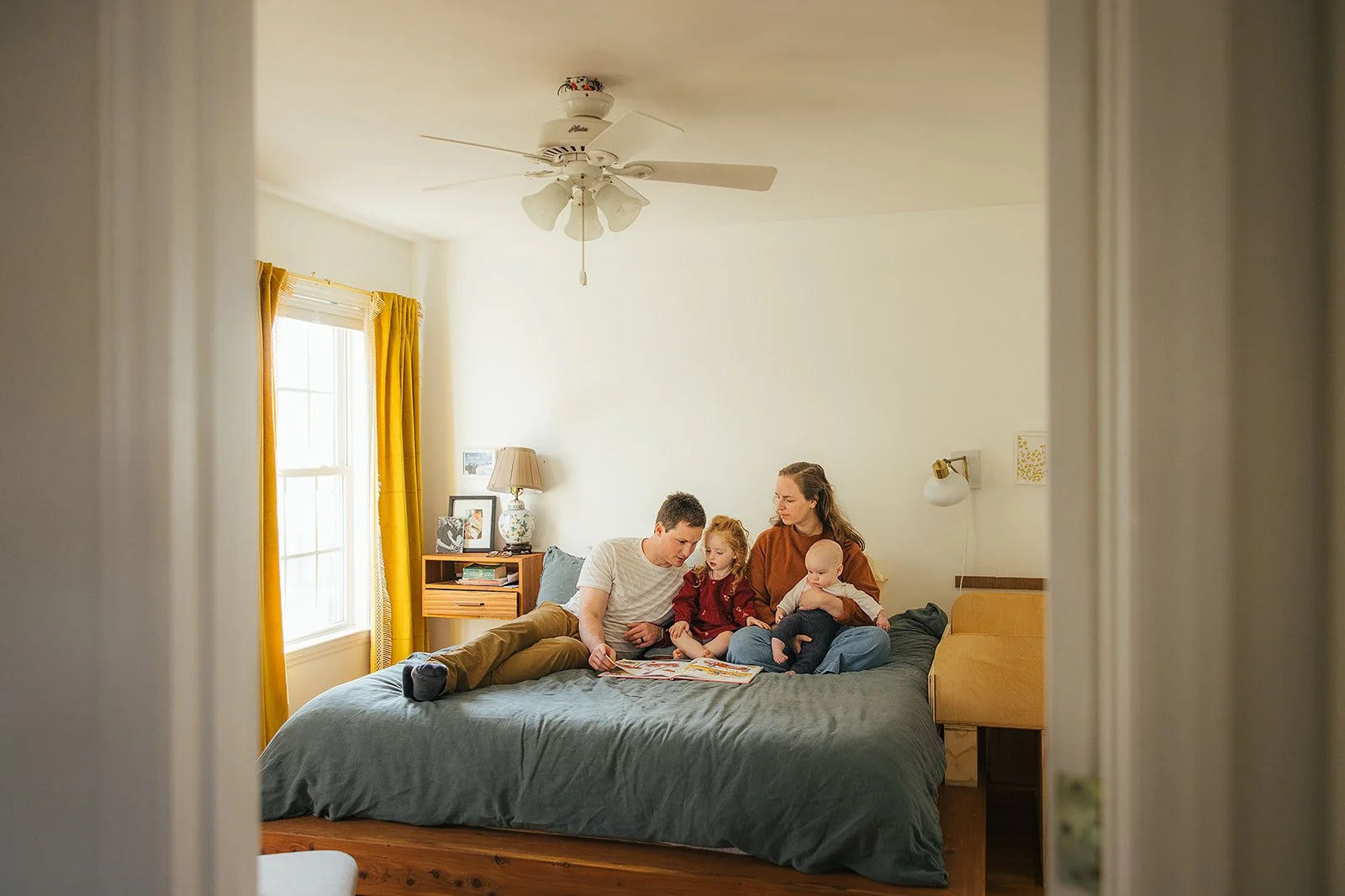 Family of four reads together on bed captured through doorway during Durham lifestyle family photography session