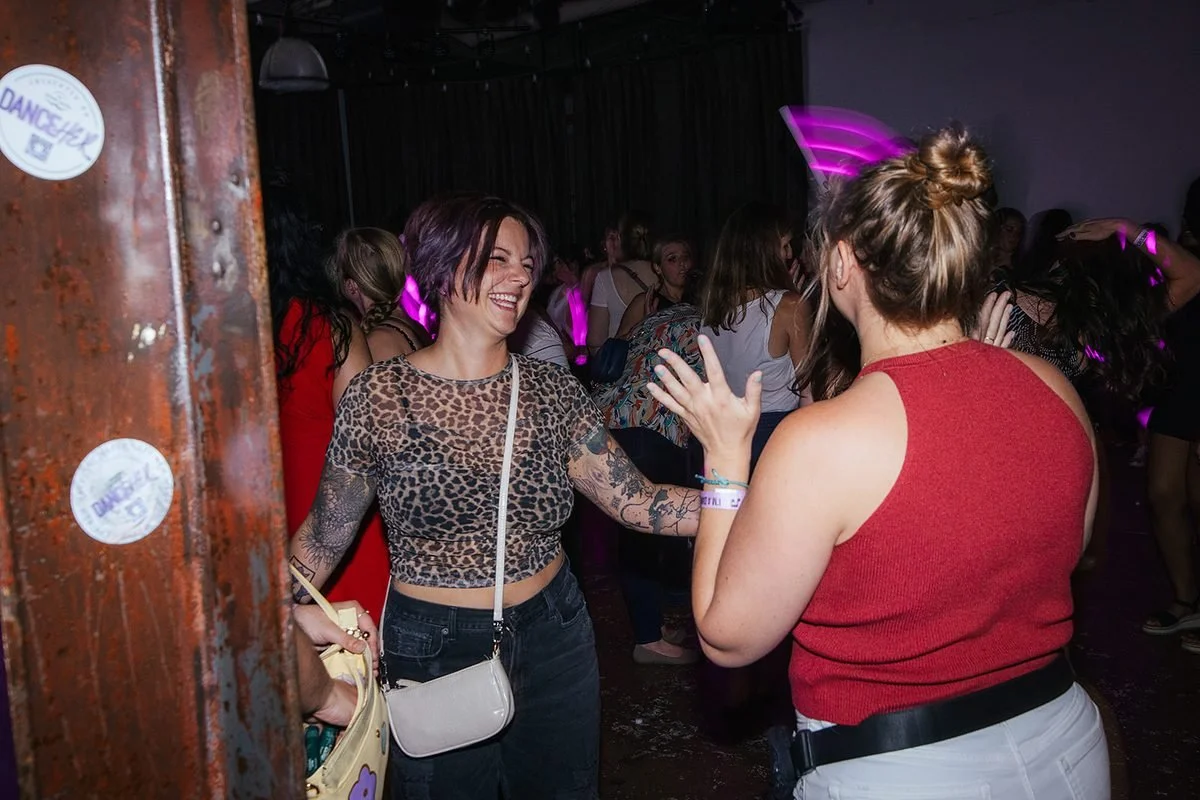Two women laughing and greeting each other near the entrance of the Dance Her event at Transfer Co, Raleigh NC — a warm, real moment