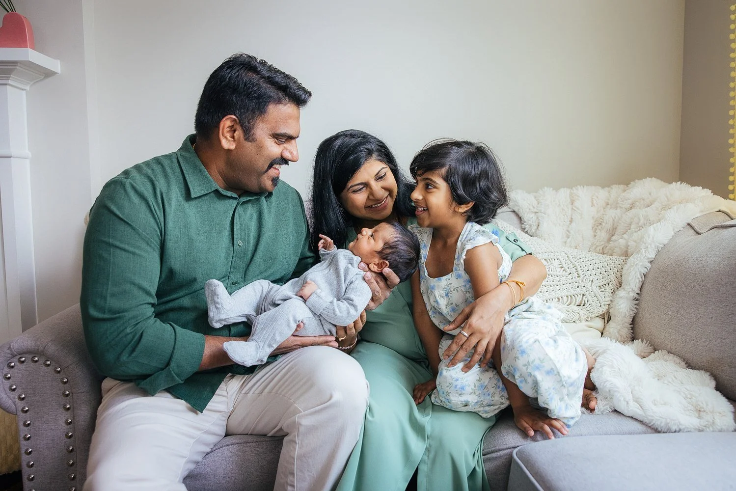 Mother, father, newborn baby, and older daughter share a warm candid moment on the sofa — lifestyle in-home newborn family session in Apex, NC.