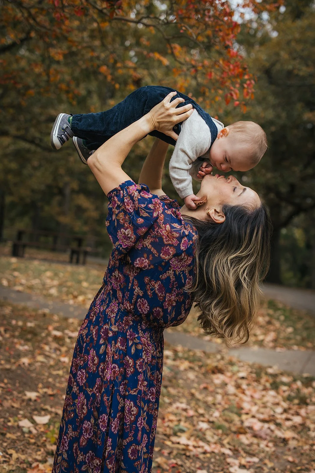 Mom lifts baby boy for a sweet nose - to - nose kiss against fall tree canopy at Duke Park during Durham NC motherhood photography session