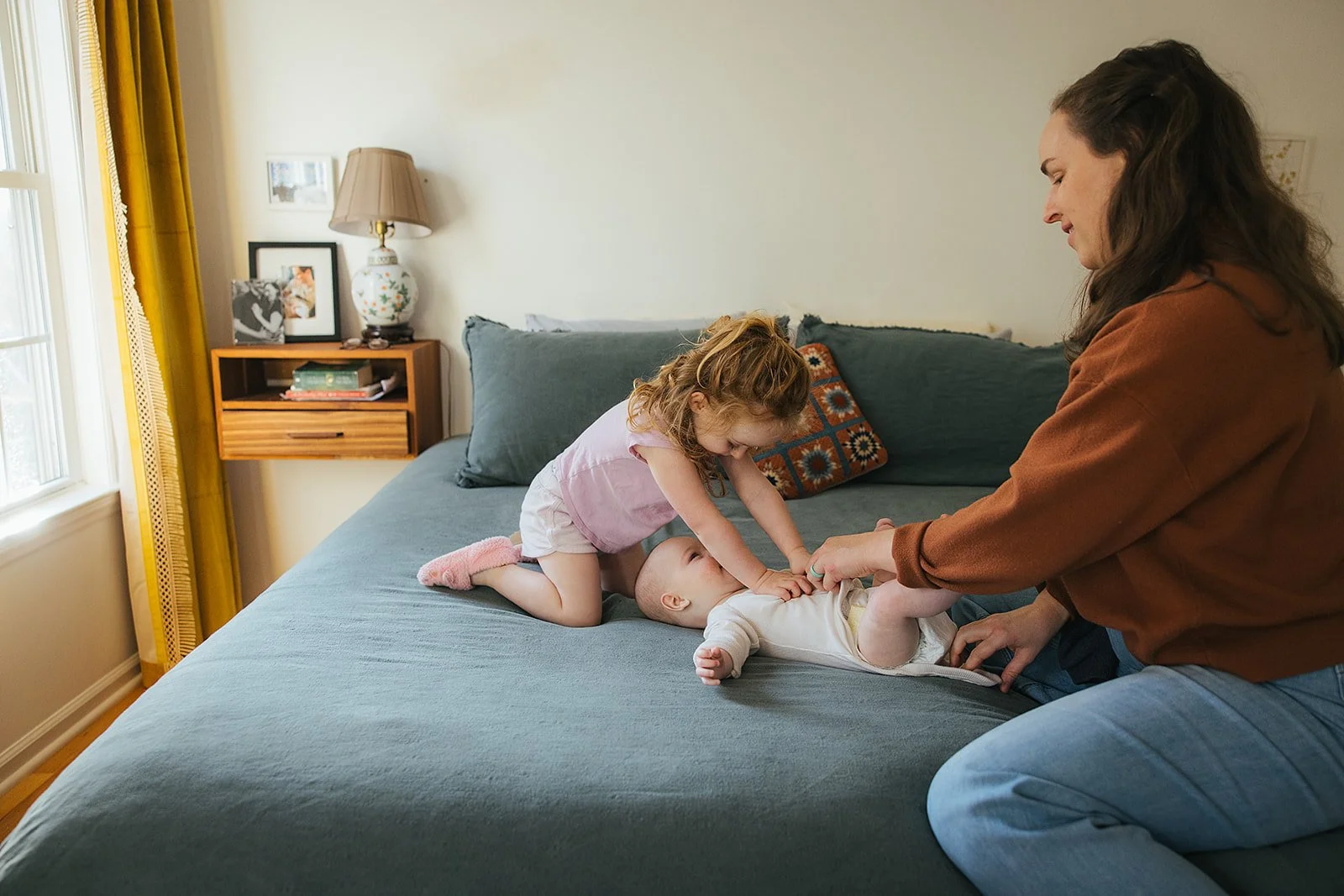 Big sister leans in to kiss baby sibling on bed during Durham in-home lifestyle family photography session